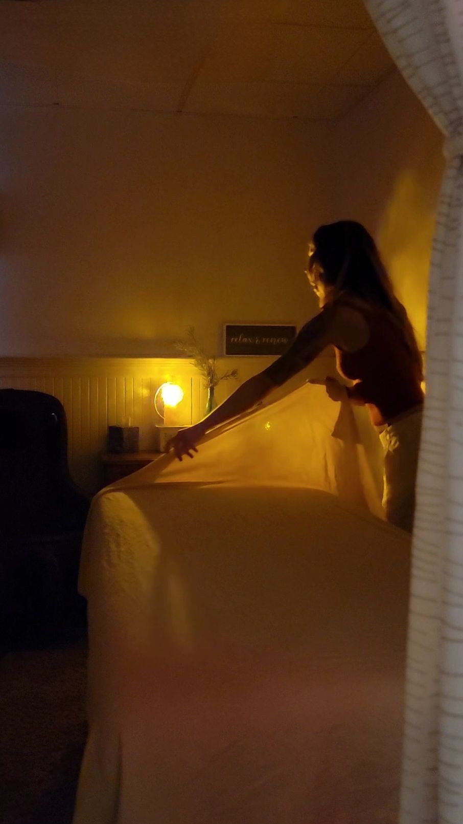 Person arranging a bed in a dimly lit room with a glowing lamp on the nightstand.